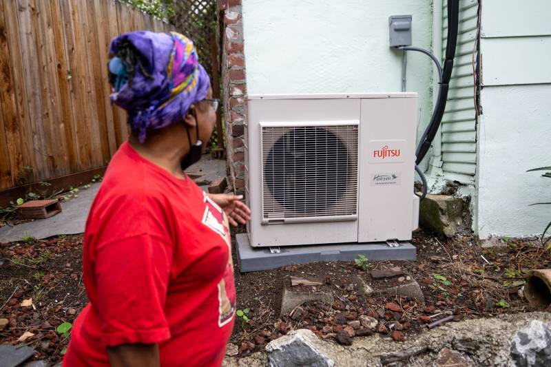 Woman in red shirt points to metal heat pump that sits along the outside of her home.