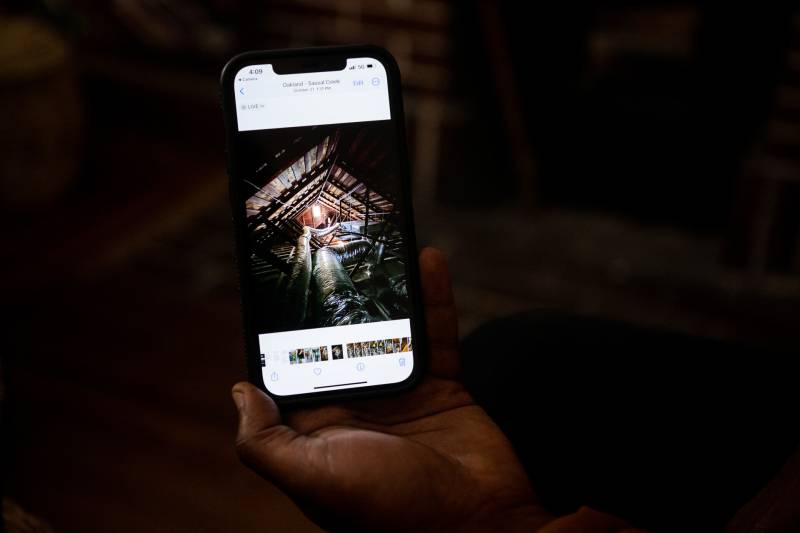 A hand holds an iPhone showing an image of an attic with silver ductwork running along the floor.