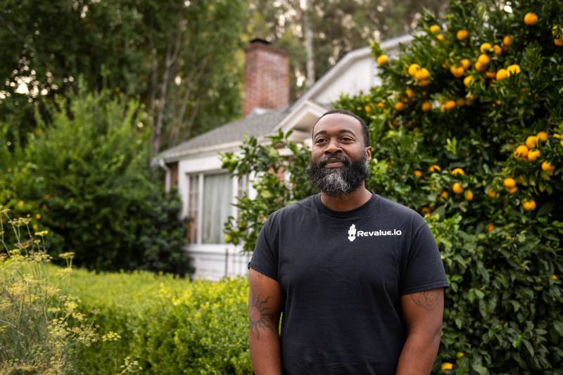 Black man in black shirt with white letters that say "revalue.io" looks towards the camera while standing in front of a home and citrus tree.