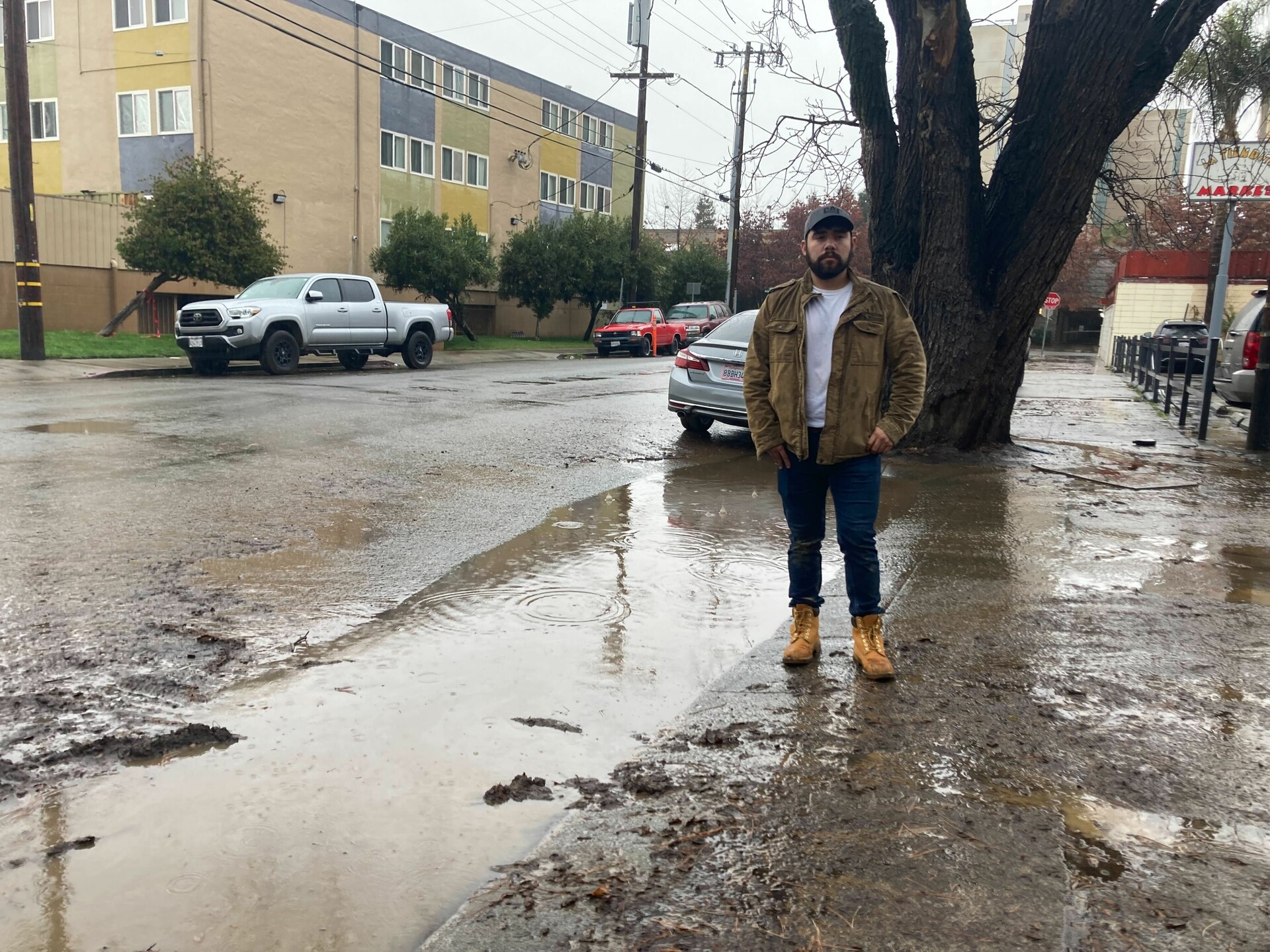 A man in a brown jacket and yellow boots poses by muddy water. 