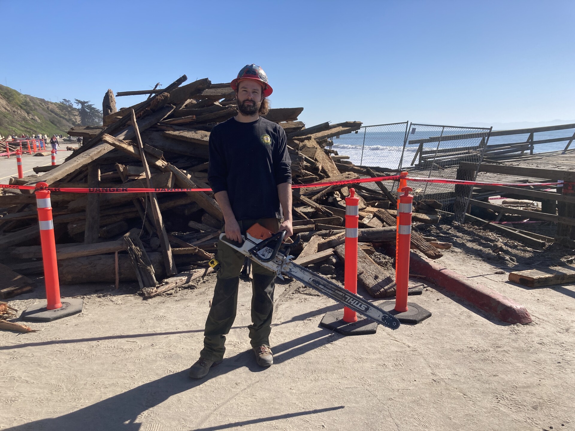 A bearded man with a chainsaw stands in front of piles of wood. 