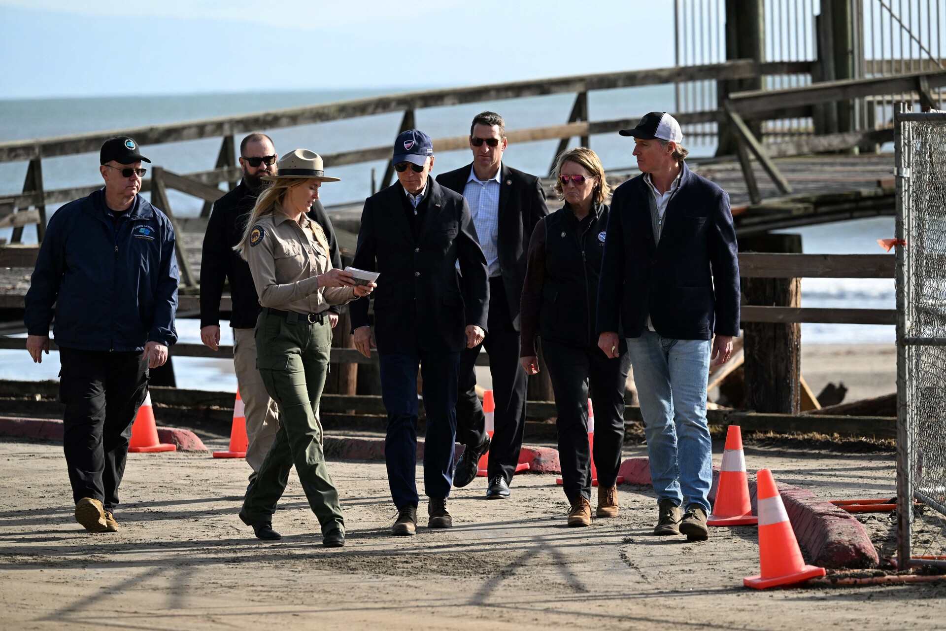 Government officials in dark coats walk in front of a broken pier. 
