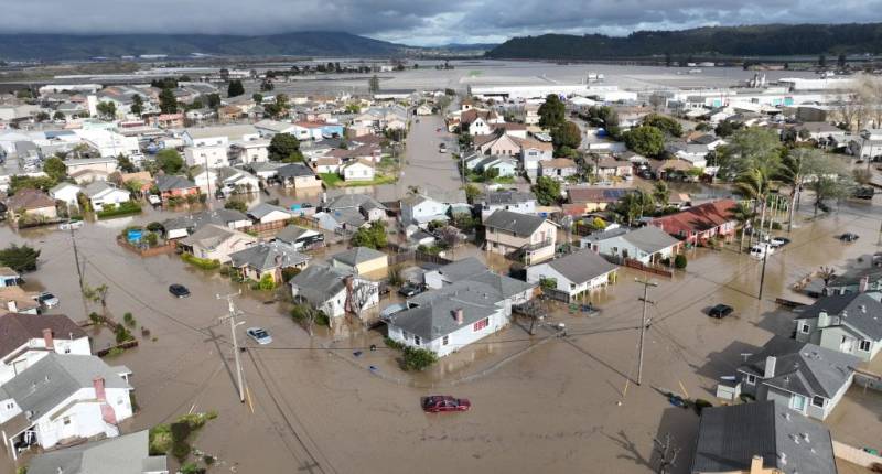 A group of houses, streets and cars submerged in brown muddy water. Dark skies and mountains are in the background.