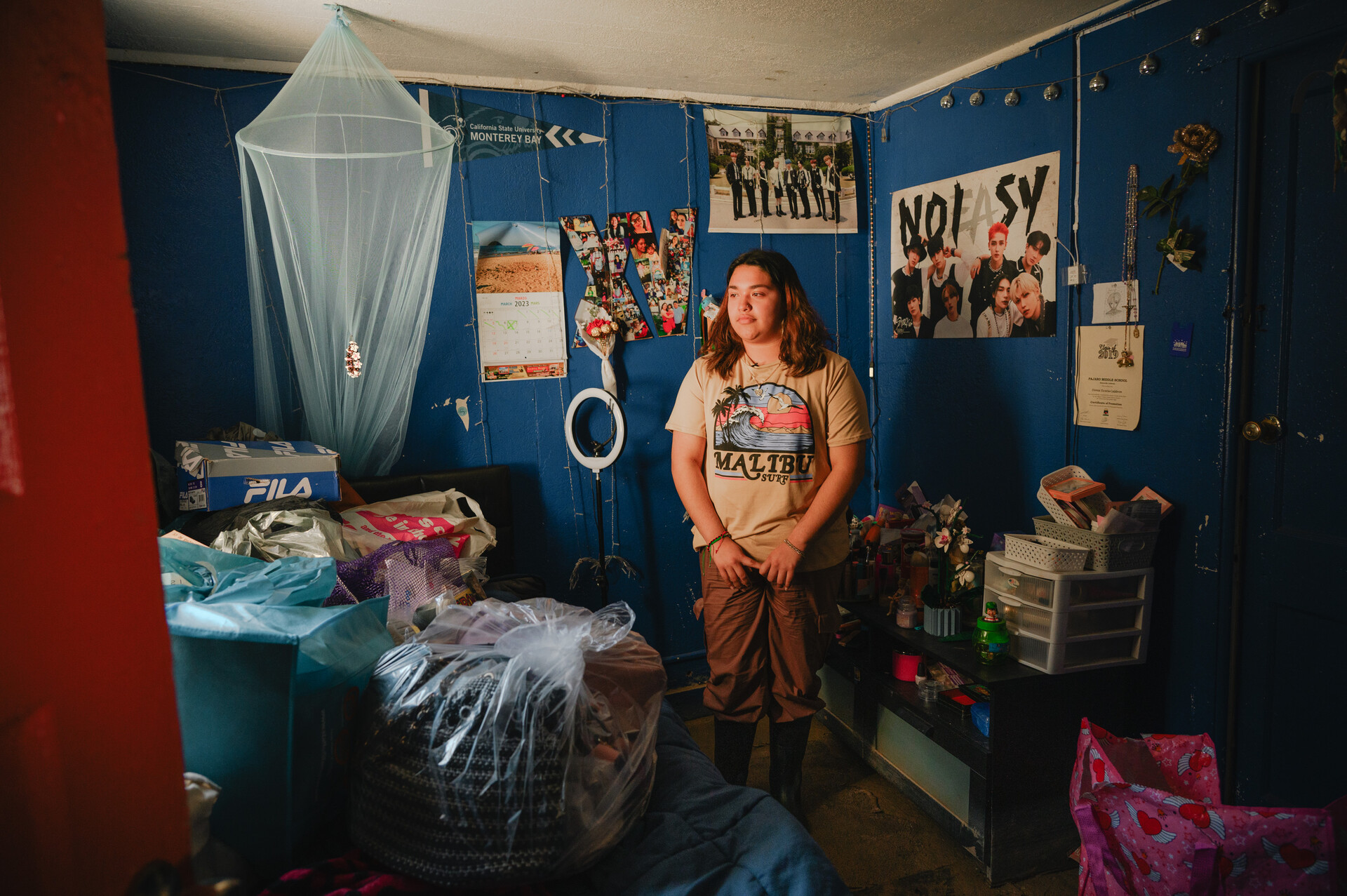 A young person in rubber boots stands in a small bedroom with posters on the blue-painted wall and mud on the floor.