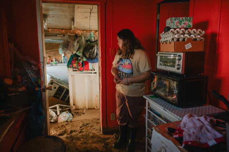 A young person wearing rubber boots stands in a small galley kitchen, where appliances on piled on the counter, looking at a mud-caked floor.