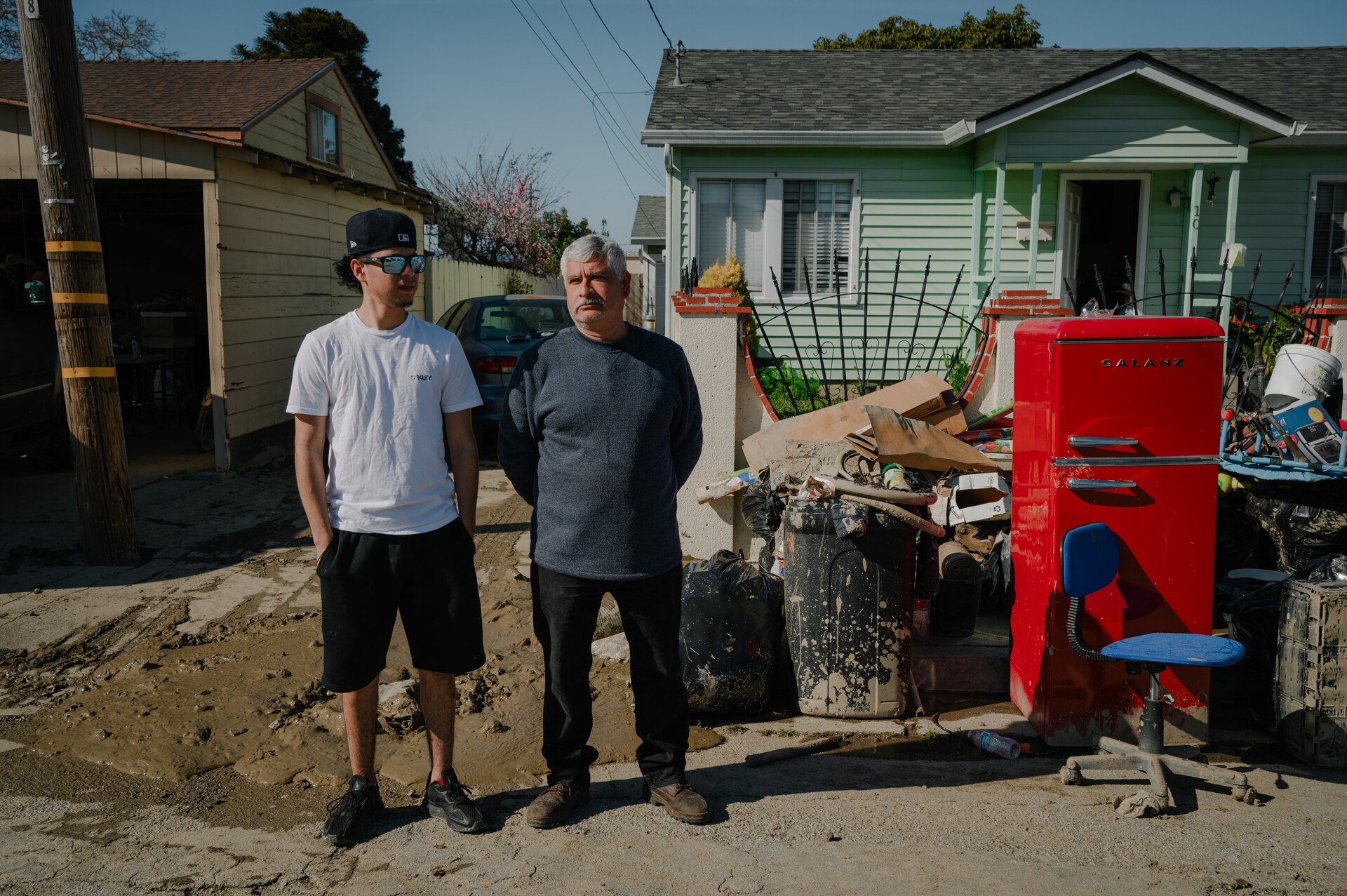 An older man and younger man stand outside a light-green house, next to a pile of mud-damaged belongings, including a red refrigerator and blue office chair.