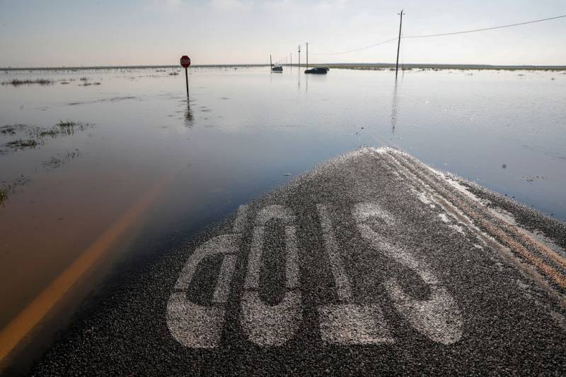 A road marked with the word STOP in white. The road is submerged in water. A car and a stop sign are enshrouded in water. A grey sky in the background.