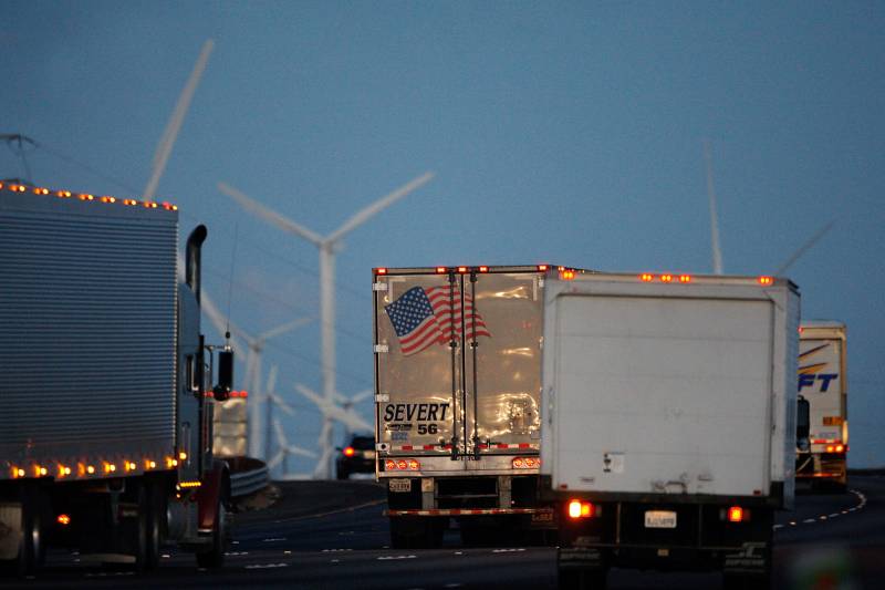 White, grey and blue semi-trucks driving along the highway at twilight. Wind turbines in the background.