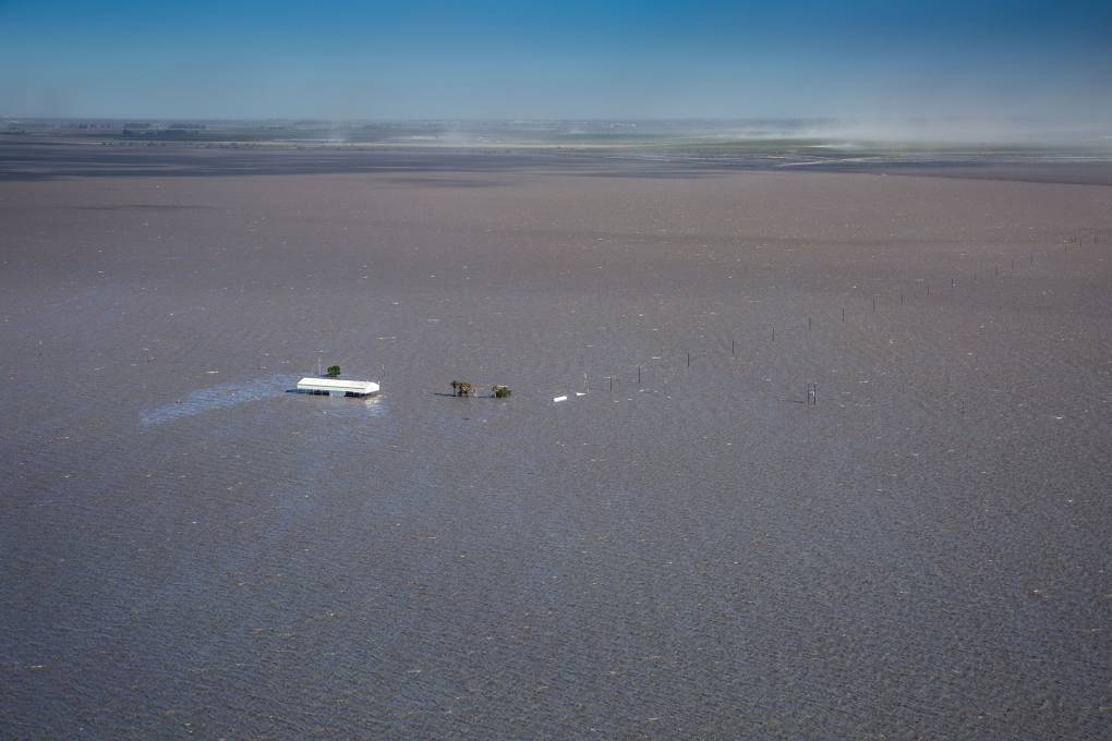 A white roof of a small home can be seen surrounded by flooded waters. The farmland where this house sits is completely flooded in dark, green waters. The tops of telephone poles stick out from the flood.