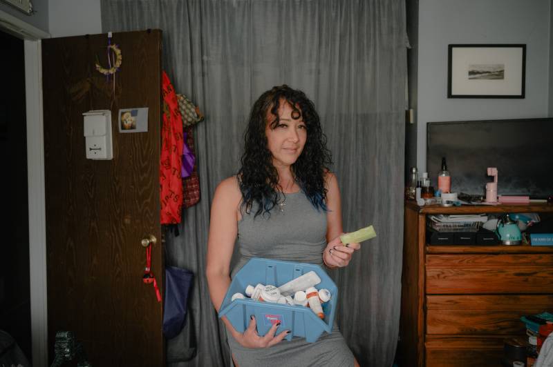 A woman wearing a grey dress holds a yellow pill box in one hand and container of prescription bottles in the other while standing in a bedroom.