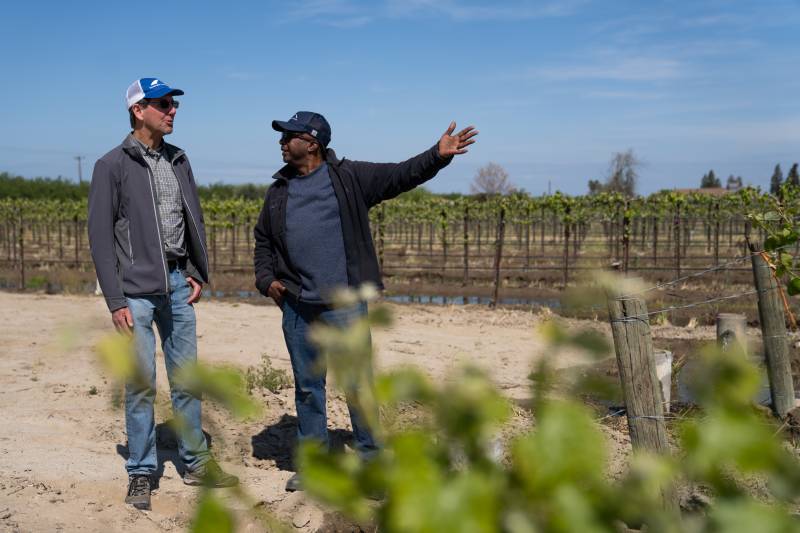 Two men, one white and one Black, both wearing baseball caps and unzipped jackets, talk to each other, one gesturing toward the vineyard they stand in with one arm raised.
