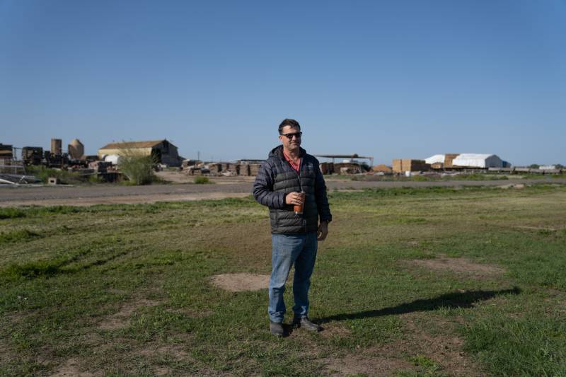 A white man with dark brown hair and black sunglasses, wearing a black puffer jacket with an orange collared shirt and jeans, stands on a farm with a to-go cup in his hand. 