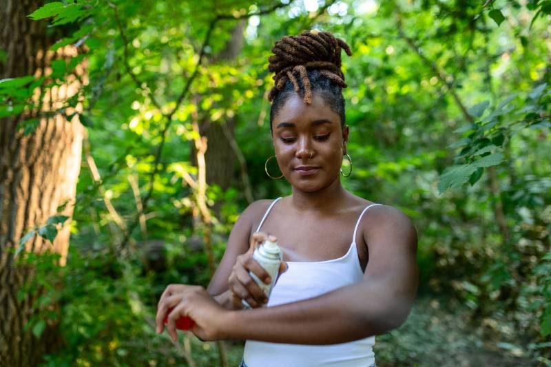 A young Black woman with locs twirled on the top of her head sprays insect repellent on her skin while in the outdoors. Background shows lush green trees.