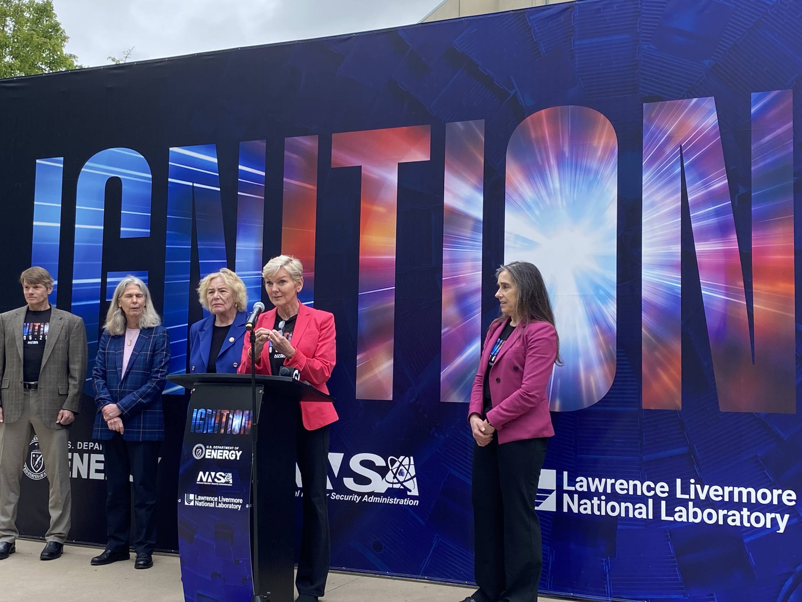 A woman with short, blonde hair and a pink blazer speaks from a podium with the words "Ignition" in big bold letters behind her printed on a banner outside of the Lawrence Livermore National Laboratory.