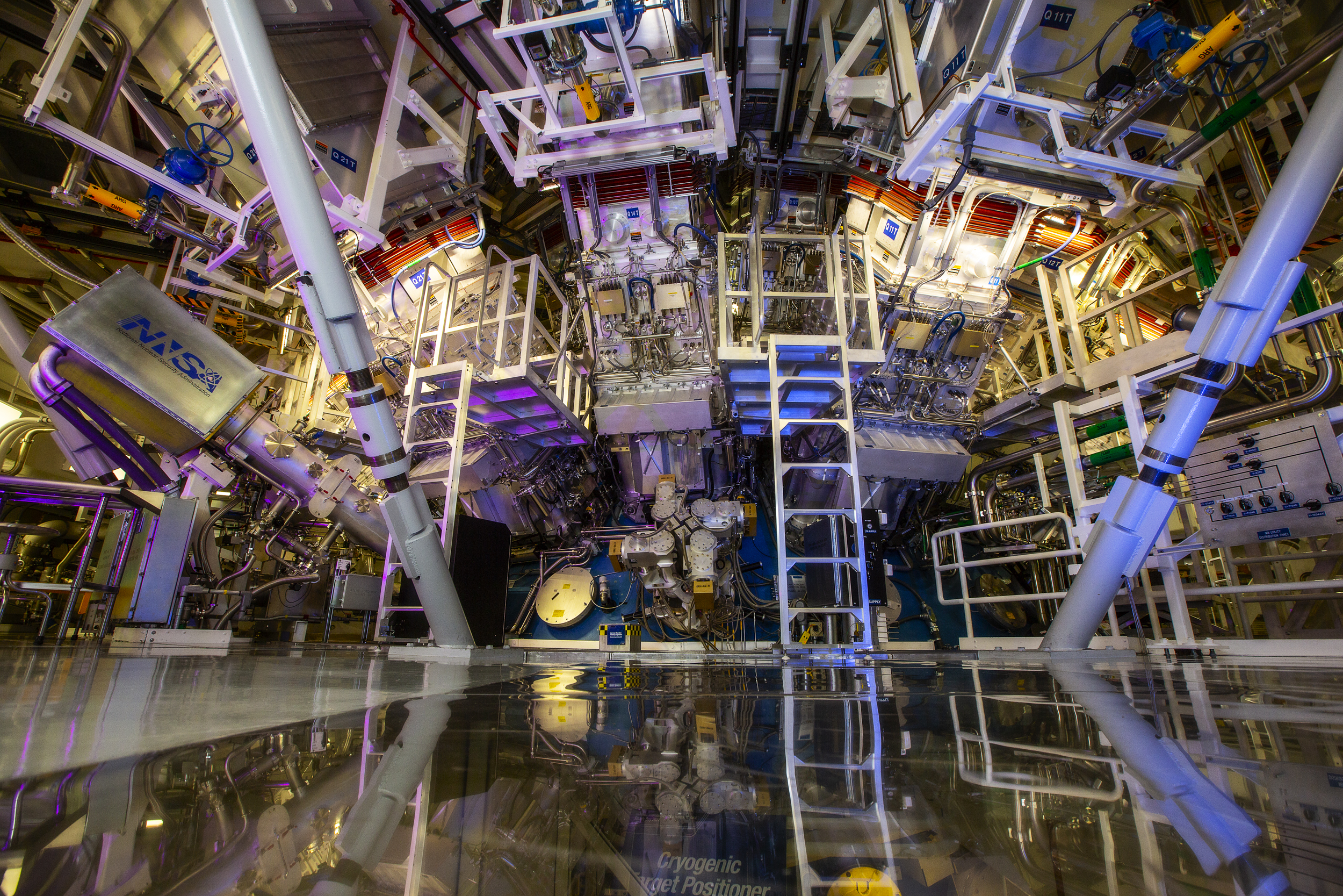 Inside a science laboratory: Gigantic metal structures with ladders leading up to various platforms. A glow of yellow and blue lights illuminate the area.