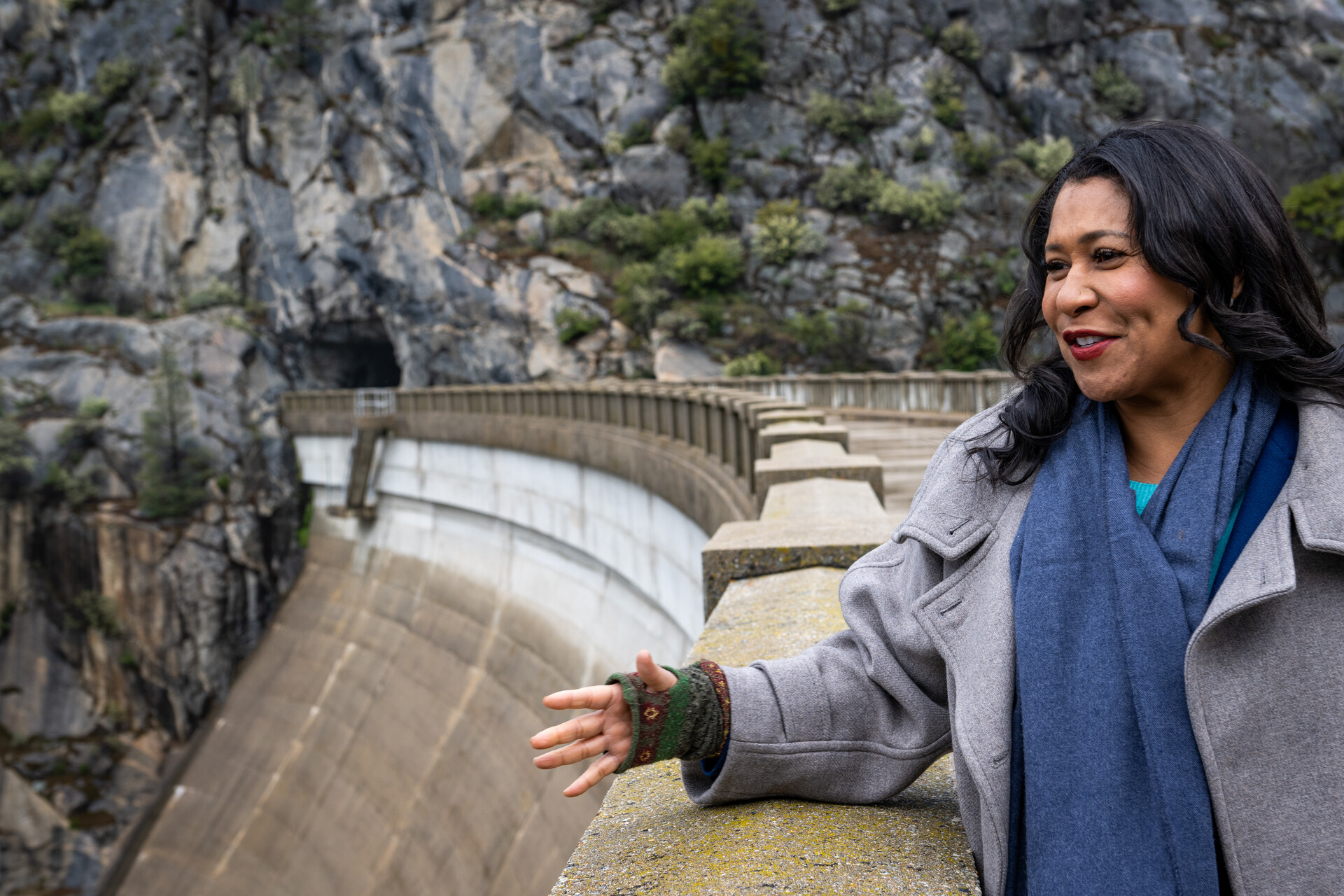 A smiling Black woman speaks while gesturing with one hand, wearing a fingerless glove and gray jacket, as the curving concrete arc of a massive dam spreads out at eye level behind her