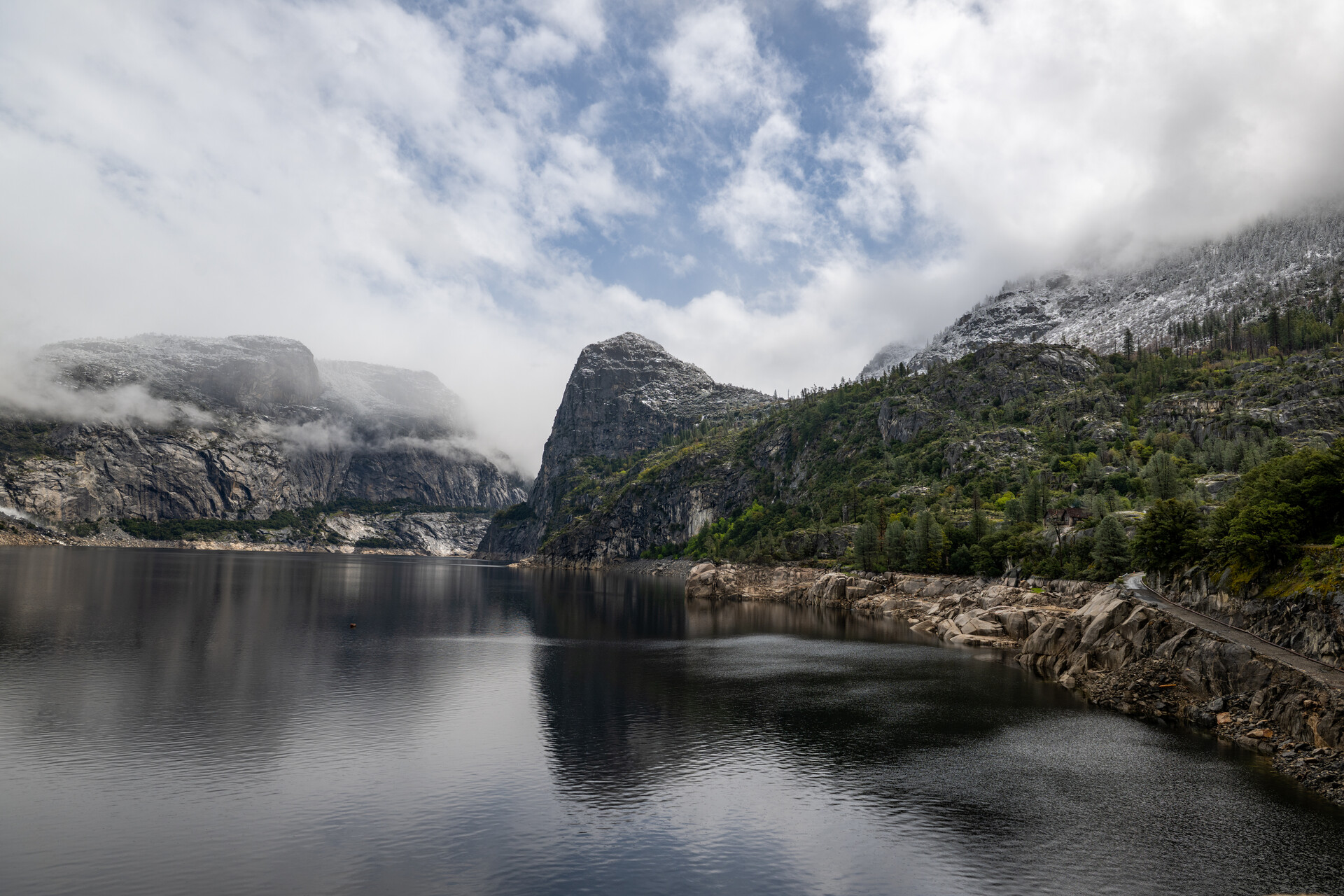 A sweeping panorama photograph shows a massive lake reflecting the shapes of steep rounded granite peaks rising above it, with alpine forest also visible