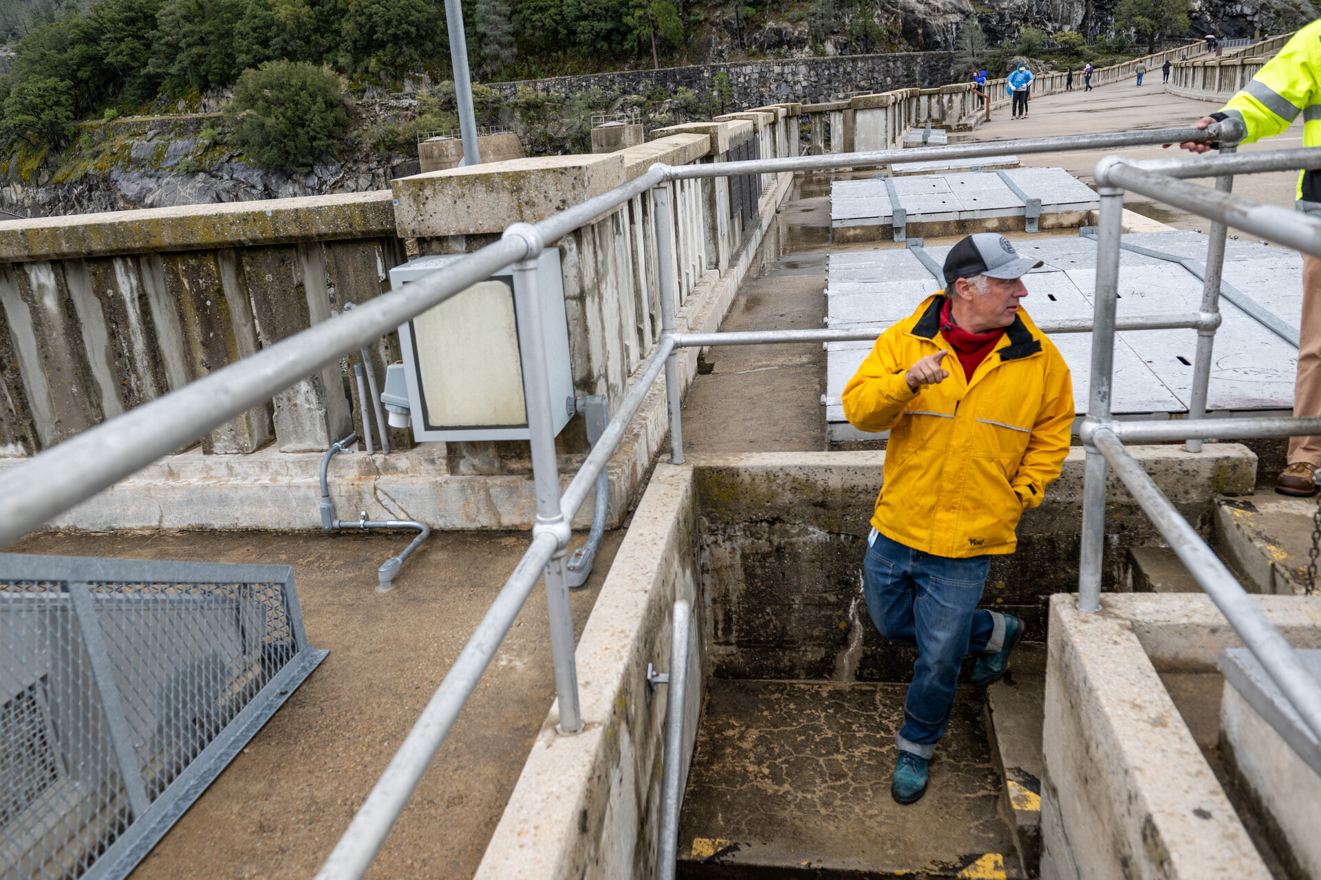 A white man in a bright yellow jacket descends concrete stairs, surrounded by metal pipes, with more concrete and a few trees visible in the far distance across the unseen water surface