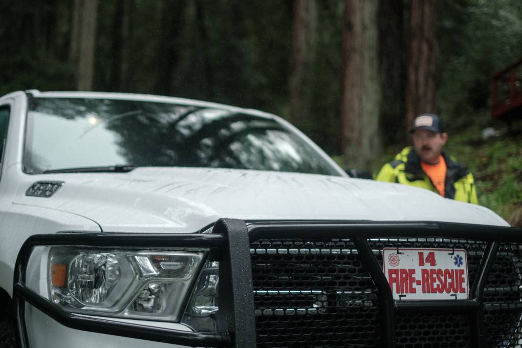 A man in a fluorescent yellow and black jacket with a black cap stands next to a white pickup truck that has a "Fire Rescue" sign on the grill.