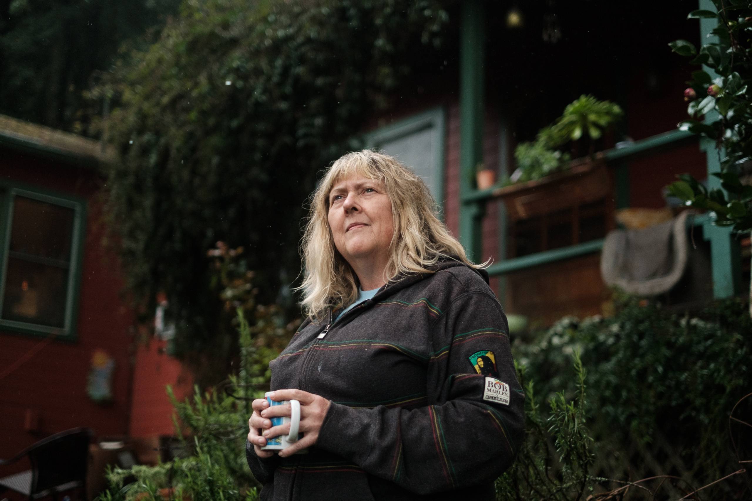 A middle aged white woman with shoulder-length blonde hair stands outside a house and looks into the distance with a mug of coffee in her hand.