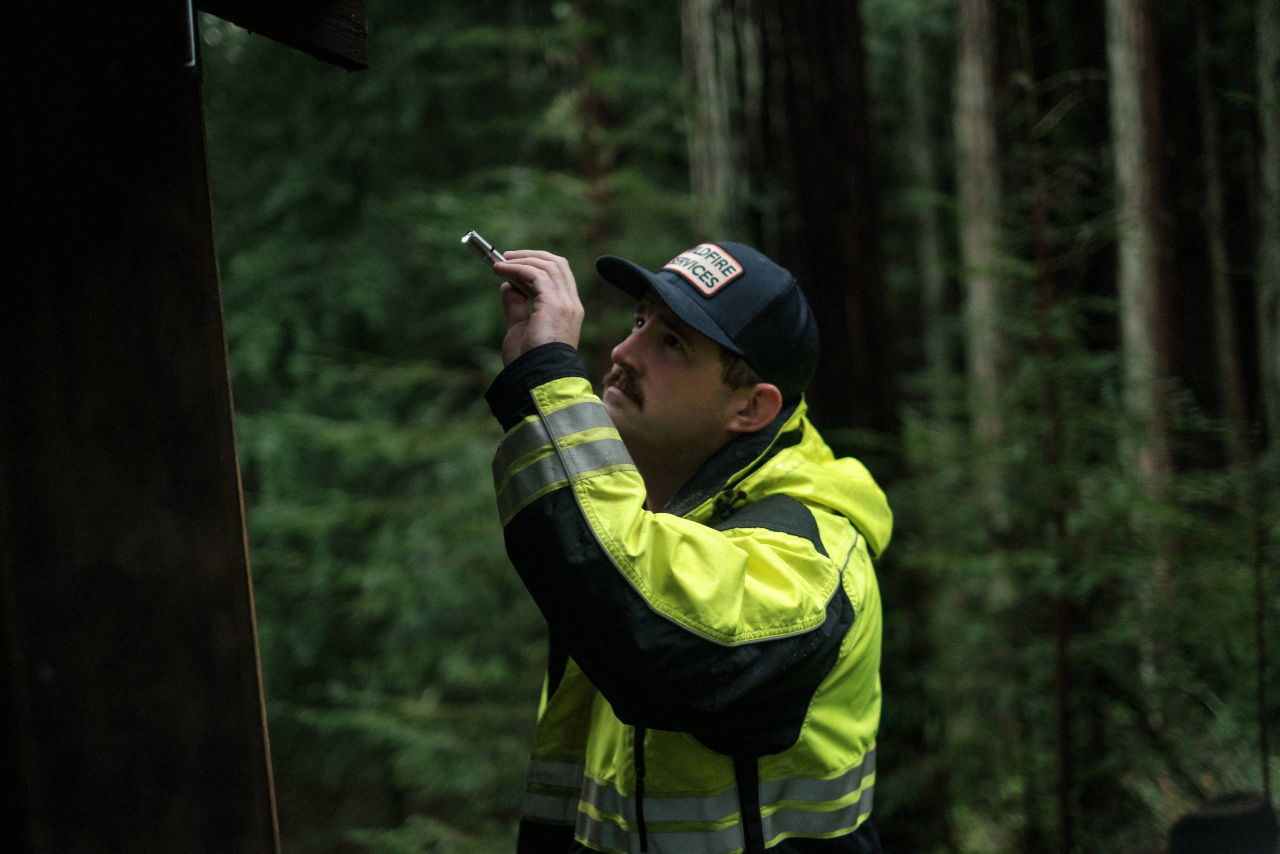A man with a cap and a fluorescent yellow and black rain jacket points a flashlight during an inspection with trees in the background.