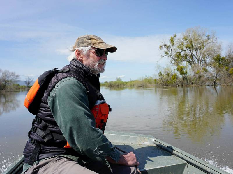 A grizzly middle-aged white man with shaggy white hair and beard, plus a camo hat and sunglasses, sits in the front of a skiff on brown water, wearing a black puffer vest and an orange life vest.