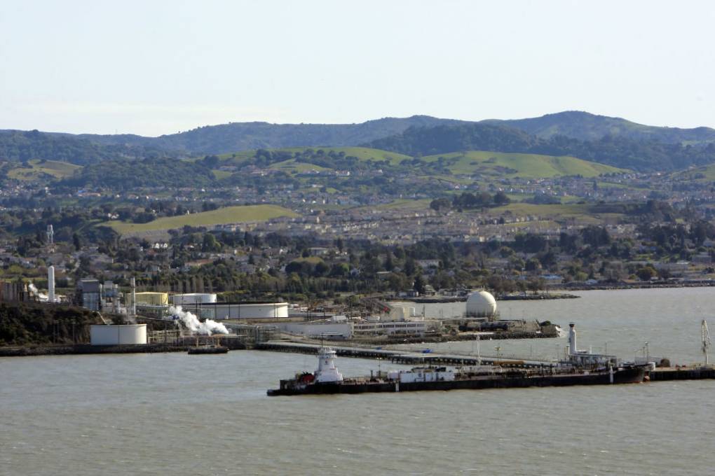 A panoramic view of the North Bay shoreline near Rodeo. In the foreground are oil refinery terminals. Behind them are houses and hills.