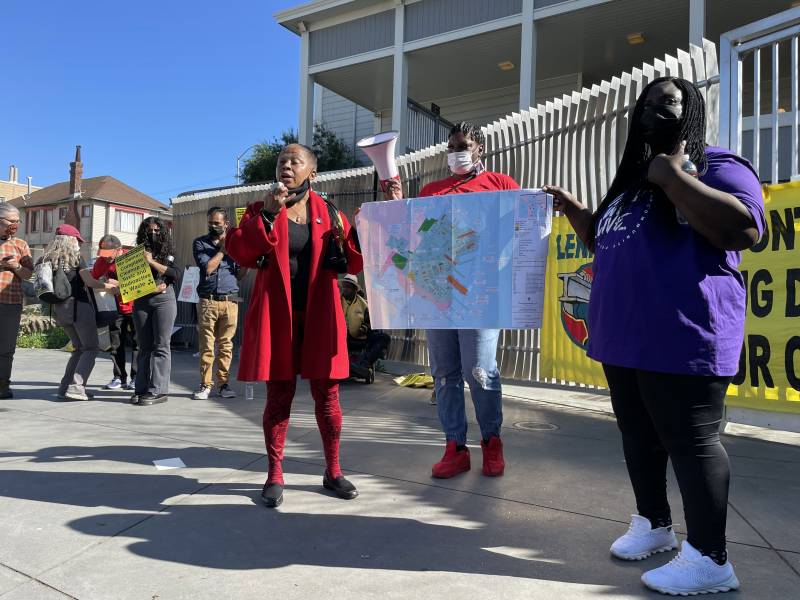 A Black woman in a red coat and red leggings speaks at a rally, holding up a map showing contaminated areas of the former Hunters Point naval shipyard. To the right of the photo, helping hold up the map, is a Black woman in black leggings, white running shoes and a purple t-shirt reading "Can We Live." In the background is a group of Black people attending the rally, dressed in denim or khaki pants and a range of colors of shirts, from blue to red plaid. The rally is in front of a tall white fence.