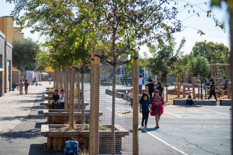 Two children are seen walking next to a tree line outdoors in the schoolyard.