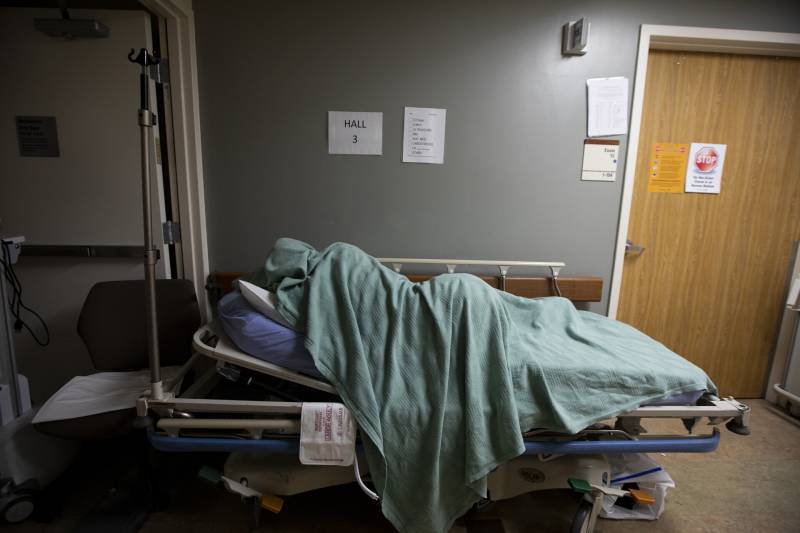A person lays in a hallway in a medical bed with a green sheet draped over them.