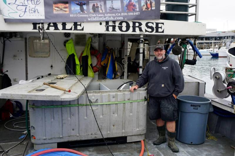 A fisherman wearing a black baseball cap, black sweater, and black shorts is standing at a fishery posing for a photo. There is a large blue bin next to him on the far right of the photo. The fisherman's right hand is rested on a large white compartment that is half opened. 
