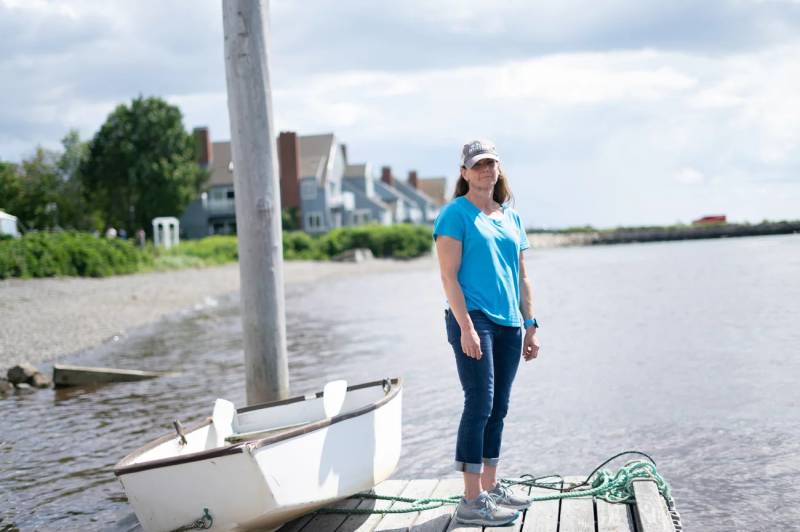 A white woman in a gray baseball cap, light blue t-shirt, blue jeans, and gray sneakers stands on a wooden surface next to a white boat. The backdrop is a lakeside with houses in front of the lake.