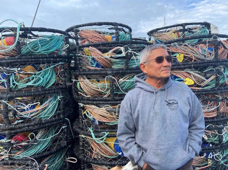 A fisherman with gray hair is wearing a gray sweater and sunglasses poses for a photo in front of some fishing gear.