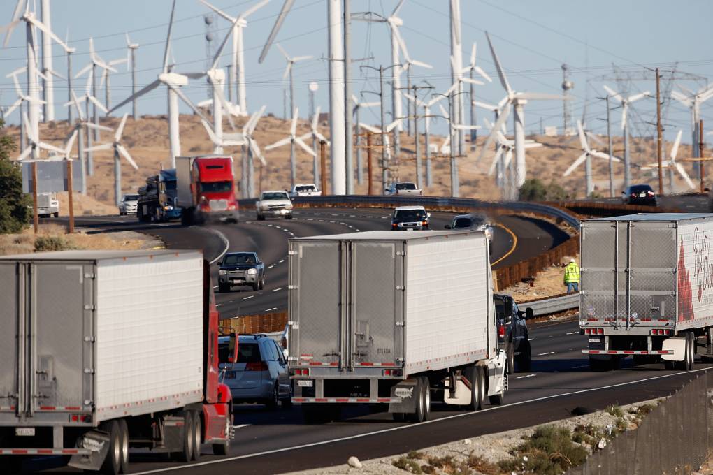 Many diesel trucks drive along a highway in California during the daytime with other family vehicles staggered in between each big rig on the road. Dozens and dozen of wind turbines are seen in the background, along with more cars, trucks and three more diesels.
