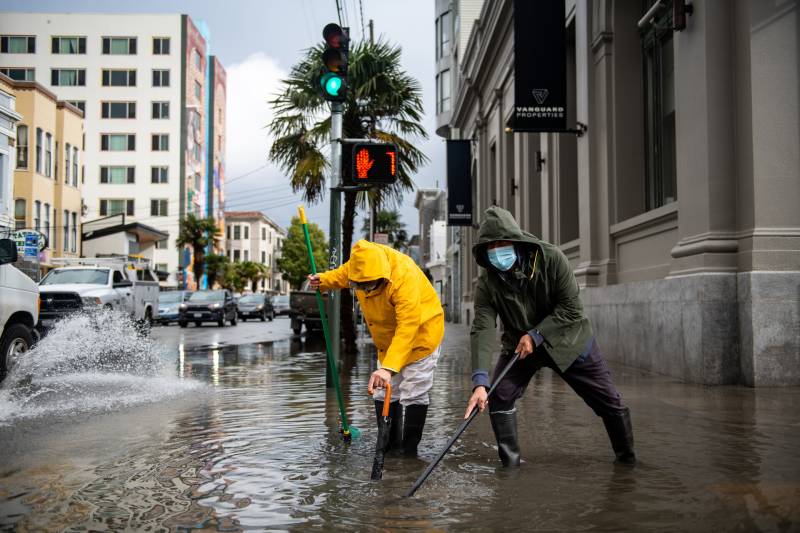 Two people in raincoats and boots use tools to try to open a drain on a flooded street.