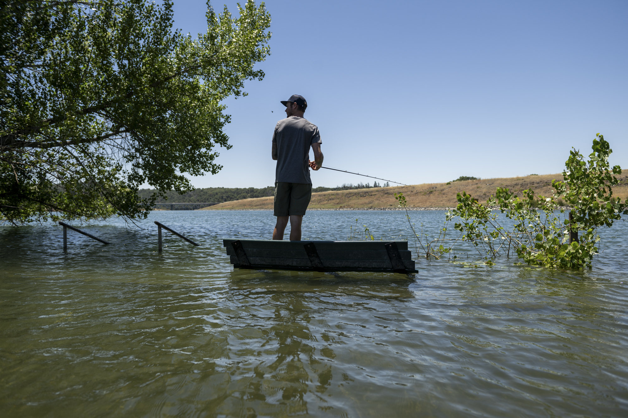 A person wearing a baseball cap wades into water holding a fishing rod in their hands.
