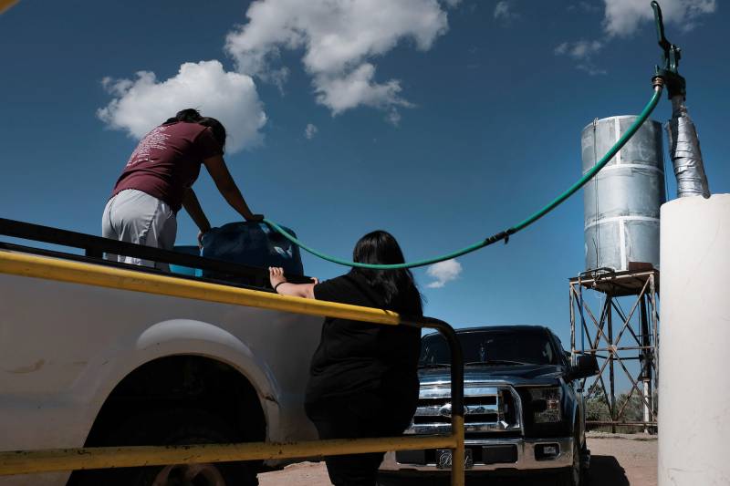 Two women are seen on a truck filling up large containers of water through a pipe. 