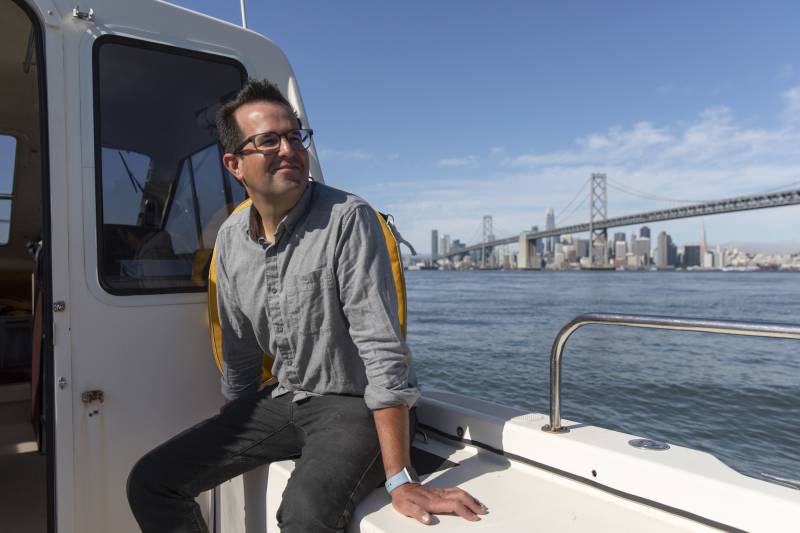 A person wearing glasses smiles while riding in a boat near a large bridge.