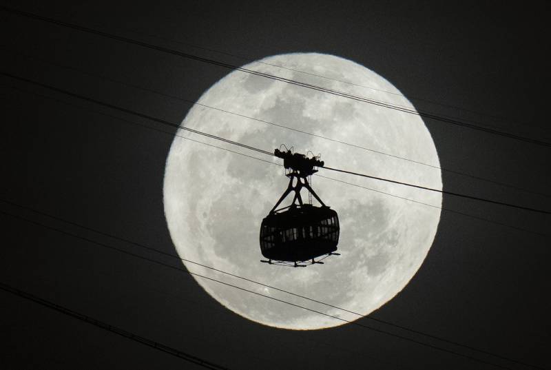 A bright white full moon is seen behind a silhouette of a cable car.