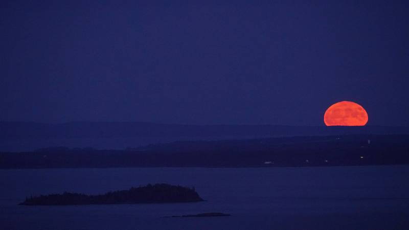 A bright orange full moon is seen in a blue background with hills and mountain silhouette. 