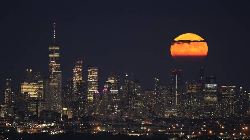 A bright full moon shines in the background of a city skyline. Clouds gather in front of the moon. 