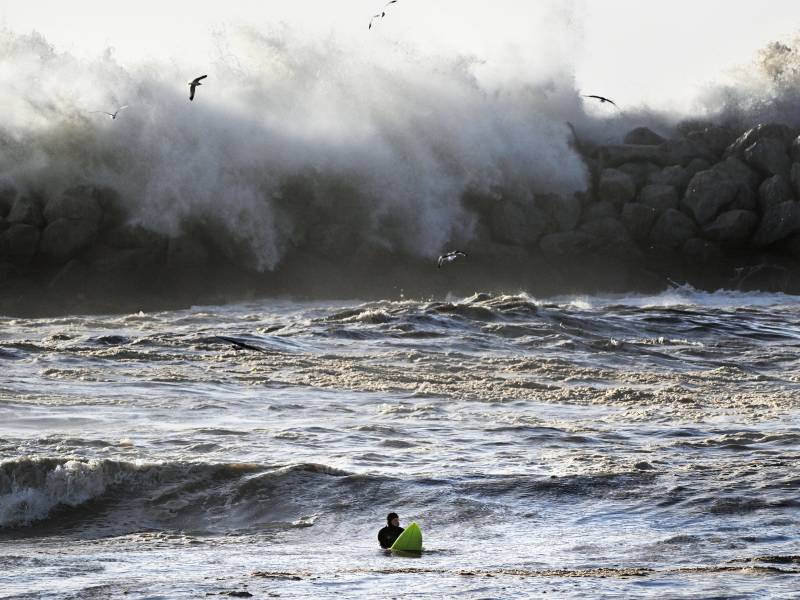 Massive waves are seen crashing against piled up rocks blocking the waves from clashing with aperson in a wetsuit, holding a neon green surf board in the water.