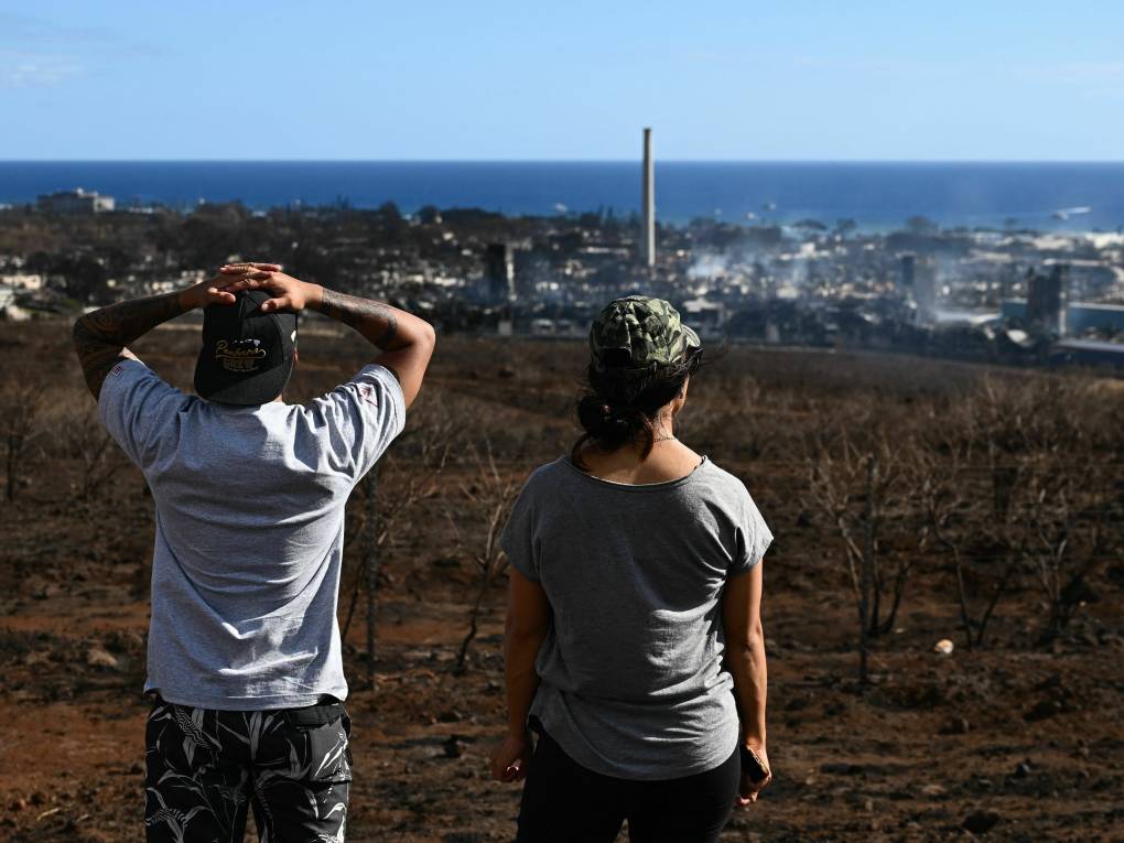 Two people look from a road above a town in the aftermath of a wildfire.