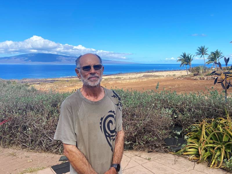 A man wearing a light brown t-shirt and sunglasses stands in front of a mountain shadowed by white clouds above it. In the background there are a few palm trees, with a view of the ocean, dry grass, and plants.