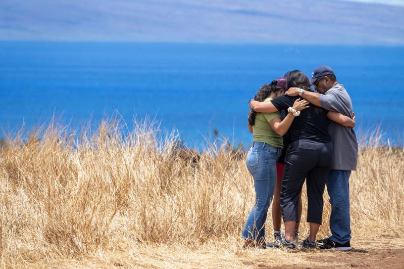 An image of three people embracing in a group hug. They are surrounded by dry grass with a backdrop of the blue ocean and skies.