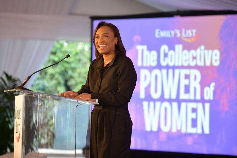 An African American woman stands at a podium wearing a full black jumpsuit with a s sign behind her that reads "EMILY's List The Collective Power of Women."