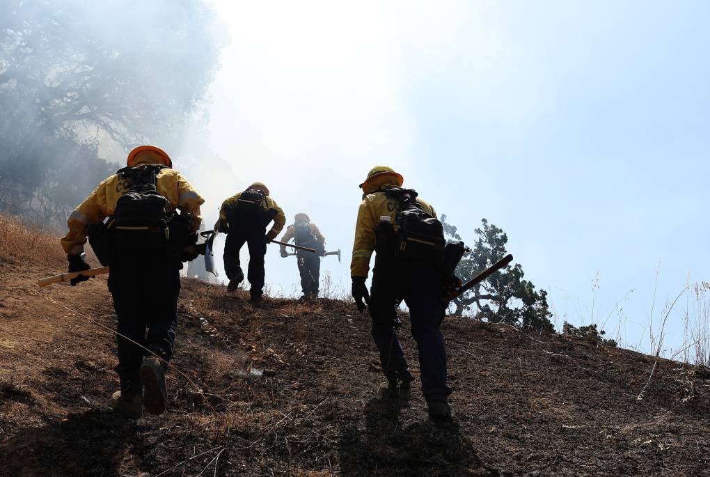 Firefighters in yellow jackets and helmets hike up a hill into a gray mist.