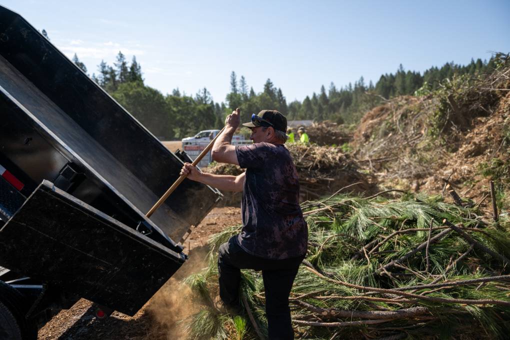 A man uses a tool to remove debris from a truck bed. Behind him is a pile of tree branches and other green waste.