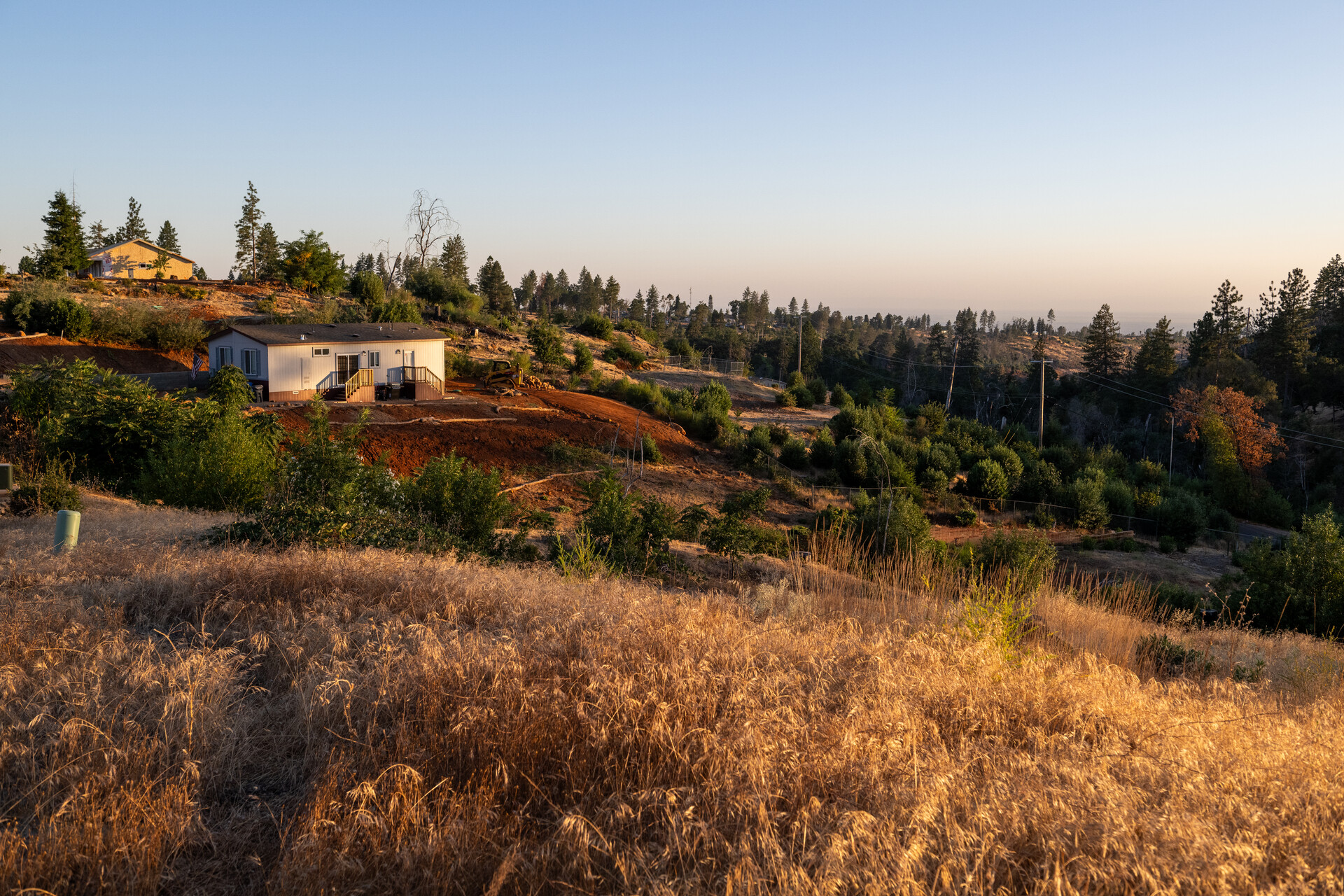 A house sits on a hillside dotted with green shrubs and dry grasses in the foreground.