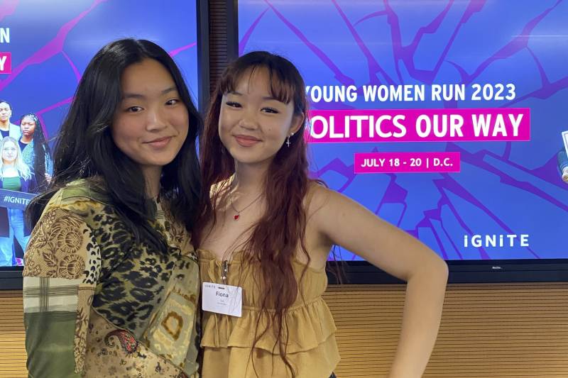 Two young Asian women stand next to each other, with the woman on the right keeping her hand on her hip behind a sign that reads "Young Women Run 2023 Politics Our Way."